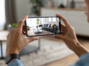 Primer plano de un teléfono moderno sostenido por manos, mostrando en la pantalla una vista de realidad aumentada con un sofá virtual superpuesto sobre una sala real desenfocada en el fondo.